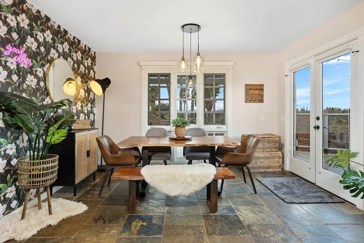 Dining room with a wooden table surrounded by seating, large windows, and a sliding door. Decor includes a floral accent wall, plants, a mirror, and pendant lights.