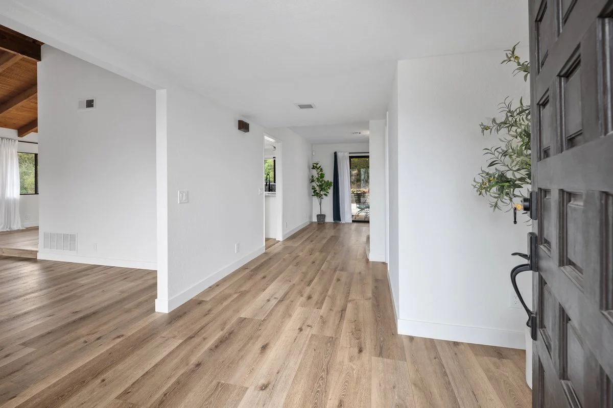 The interior of a modern house with hardwood floors, white walls, and minimal decor, viewed from the front door.