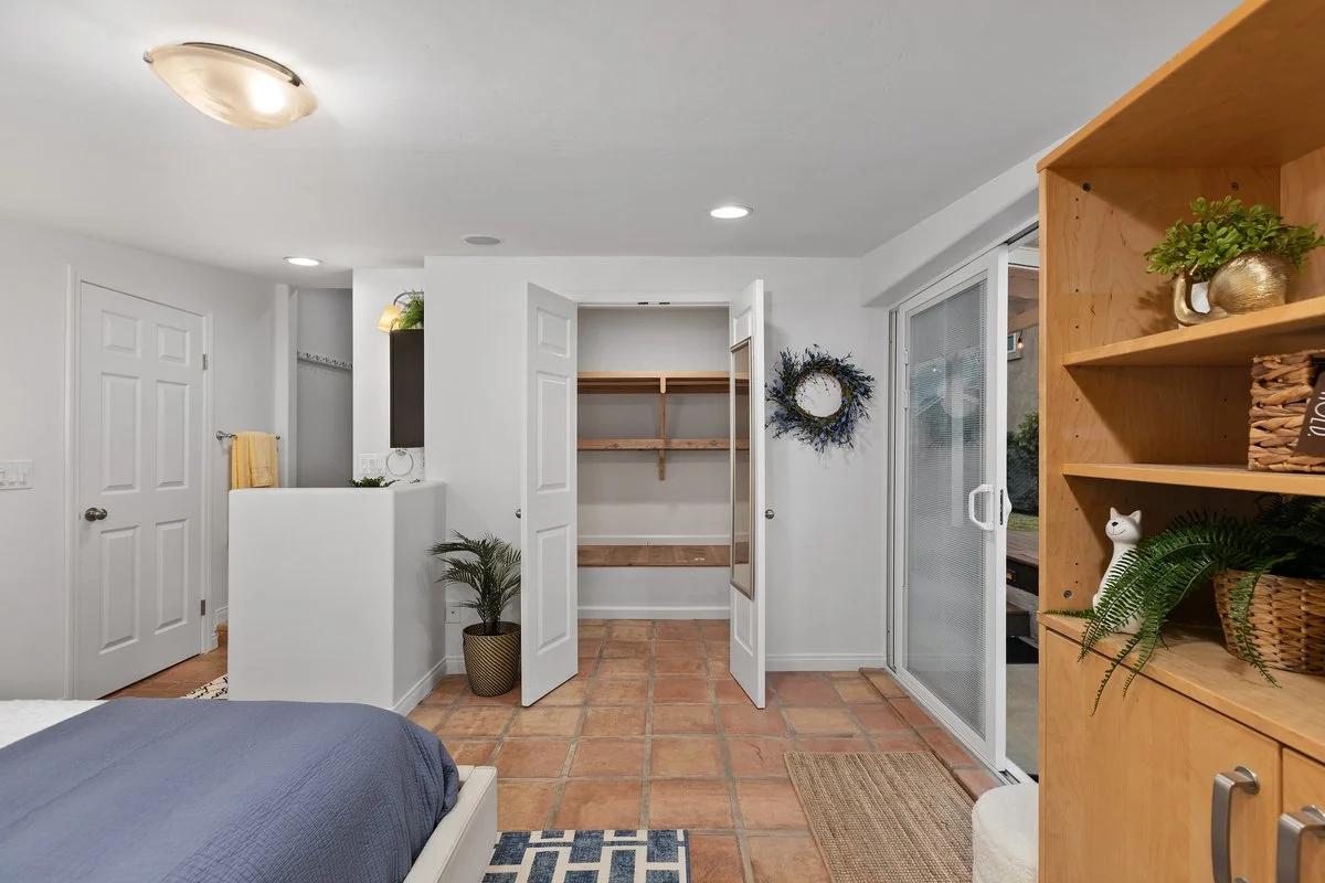 A bedroom with tile flooring, a partially visible bed with blue bedding, a wooden bookshelf with plants and decor, a sliding glass door, and an open closet.