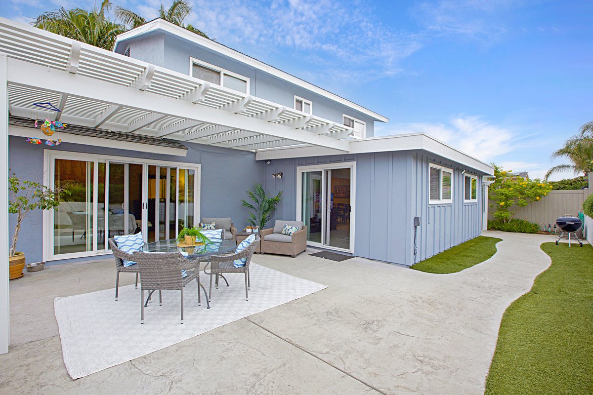 Blue house with sliding glass doors and an outdoor patio with dining table, chairs, and seating area, surrounded by greenery and blue sky.