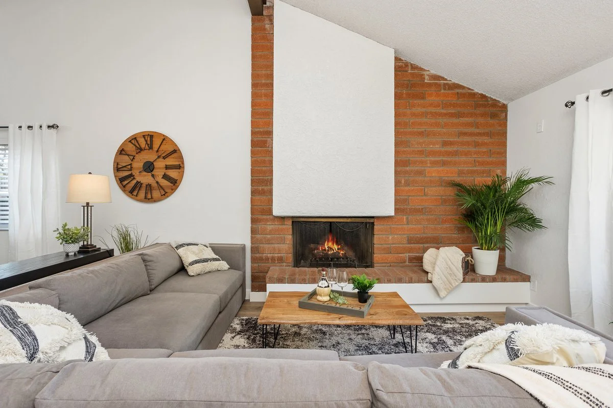 Living room with brick fireplace, beige sofas, a wooden coffee table with drinks and plants, a wall clock, and white curtains.