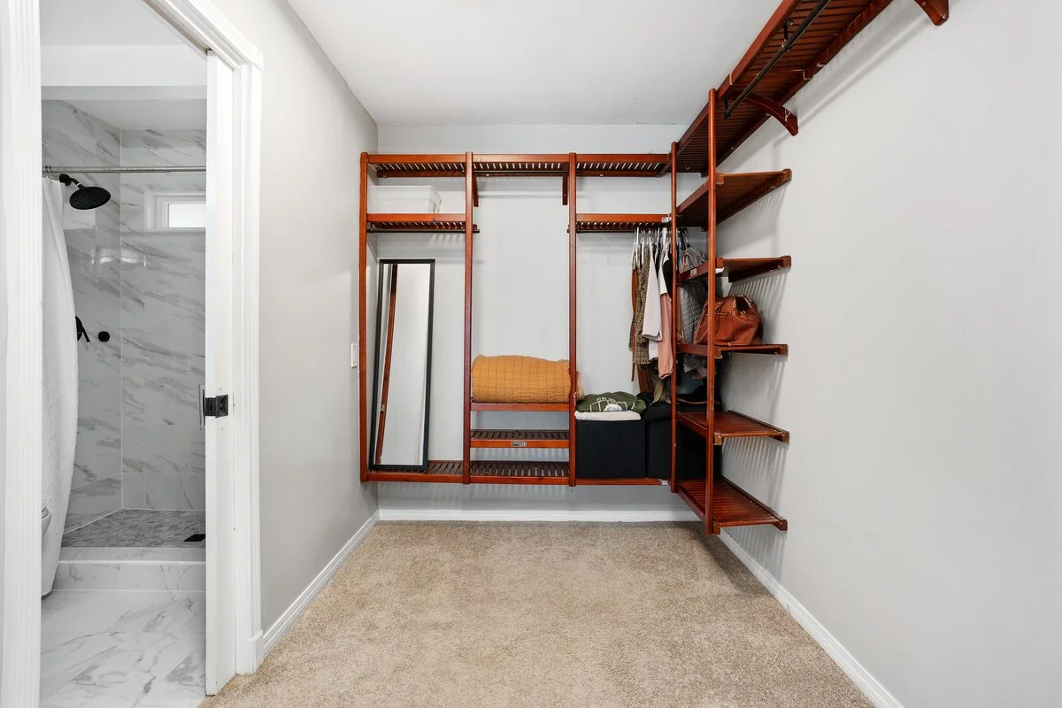 Empty walk-in closet with wooden shelves, hanging clothes, a full-length mirror, and a bathroom doorway to the left with a marble shower.