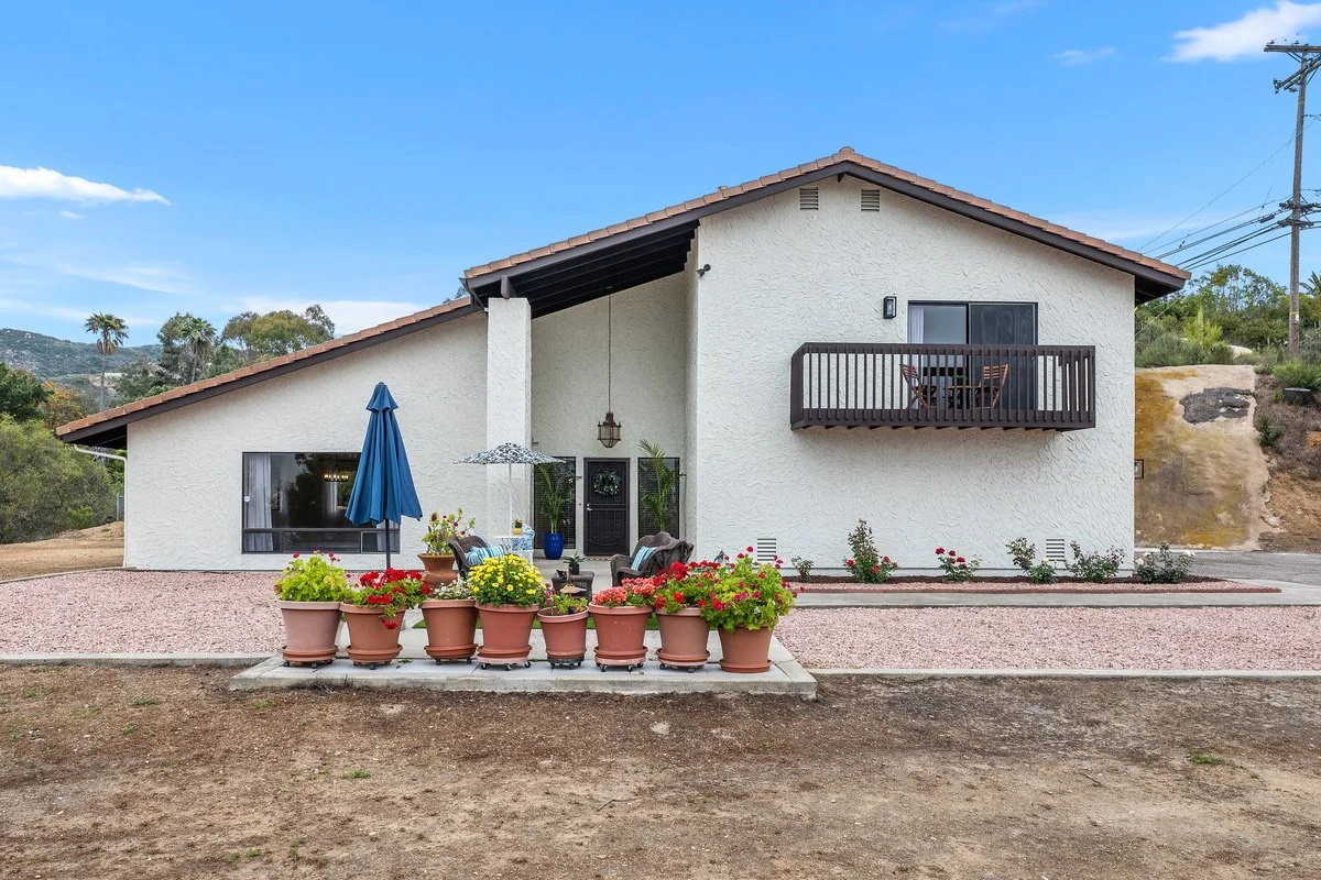 Exterior view of a modern house with white stucco walls and a sloped roof. There is a small patio with potted plants, outdoor furniture, and a large blue patio umbrella. The house has a balcony on the second floor with a table and chairs. The backgro