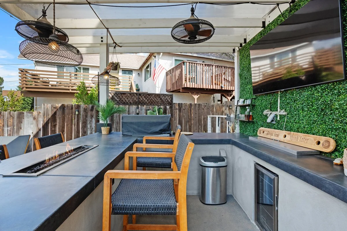 Outdoor patio with a black countertop, wooden chairs, a small fire pit, hanging outdoor fans, a large television mounted on a green wall, and a glass fireplace, set in a backyard with neighboring houses and an American flag.