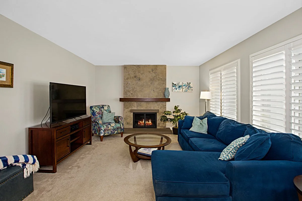 Living room with a blue sectional sofa, a glass coffee table, a floral armchair, a TV on a wooden stand, a fireplace, a potted plant, a floor lamp, and blinds covering large windows.