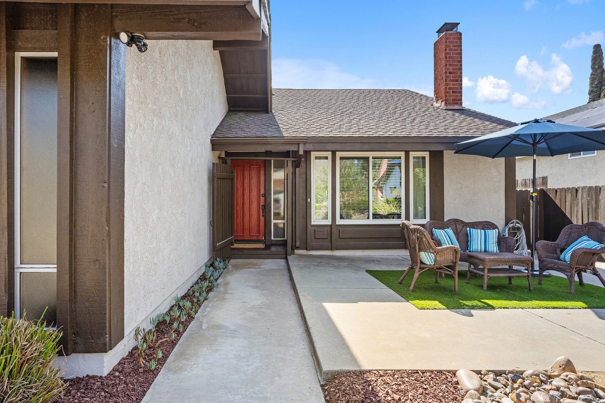 Exterior view of a house with a front porch, red front door, outdoor seating with blue-striped cushions, umbrella, and small garden area with rocks and plants.