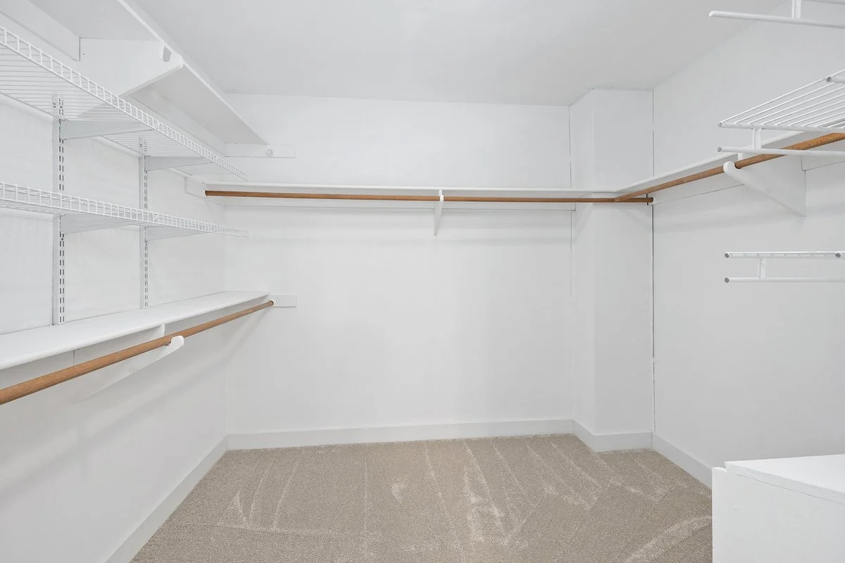 Empty walk-in closet with white walls, beige carpet, and multiple wooden and metal hanging rods.