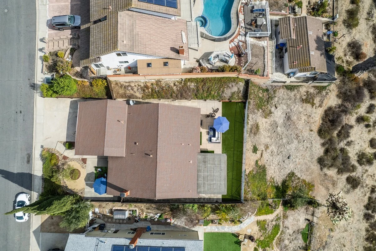 An aerial view of a residential backyard with a house, a small lawn, an outdoor seating area with umbrellas, a swimming pool, and neighboring houses, with a dirt area to the right.