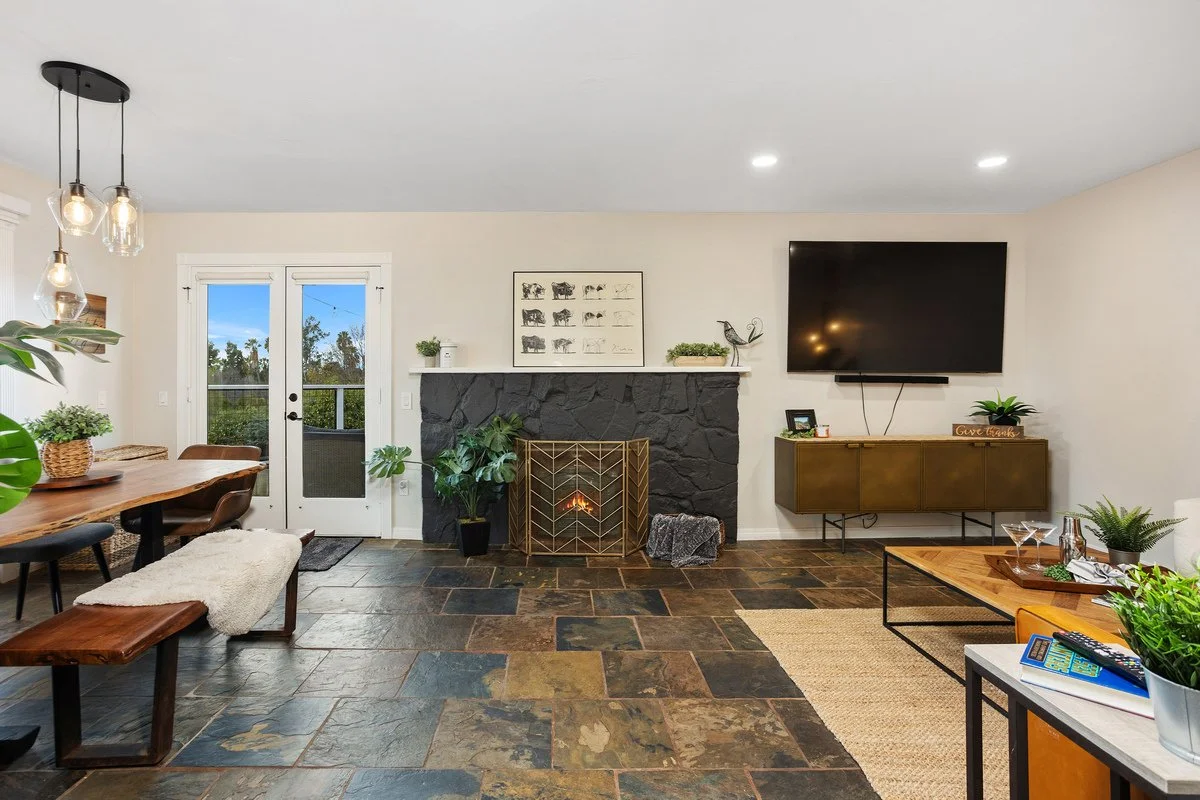 Living room with stone fireplace, mounted flat-screen TV, wooden dining table with chairs, and glass door leading outside, decorated with plants and artwork.