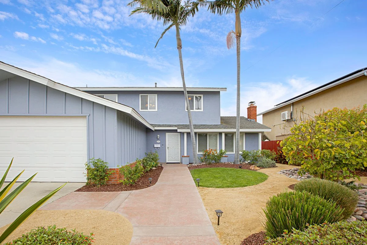 Front view of a two-story house with a gray exterior, white garage door, and landscaped yard with palm trees, bushes, and walking path.