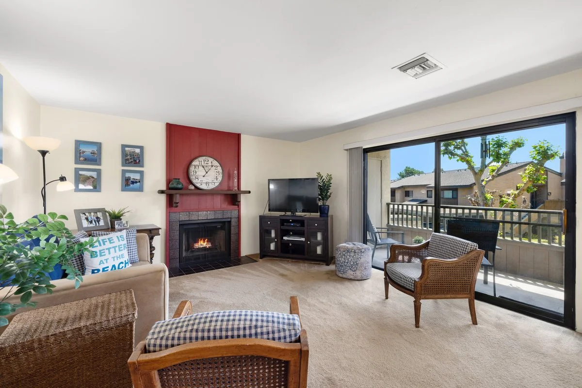 Living room with a beige sofa, wicker and upholstered chairs, a fireplace with a red accent wall, a TV on a stand, and sliding glass doors leading to a balcony with outdoor furniture and trees.