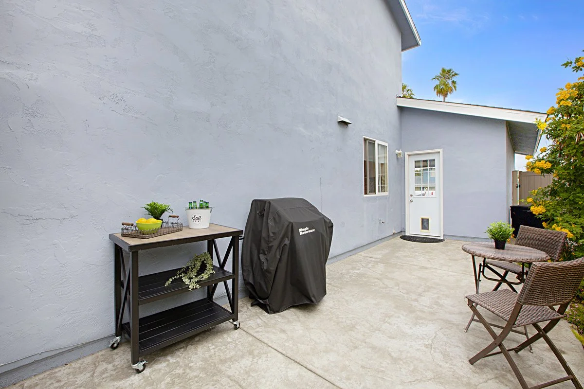 Backyard patio area with a small table, two chairs, a grill covered with a black cover, and a white door leading inside a house. There is a blue wall, yellow flowering shrub, and palm trees in the background.