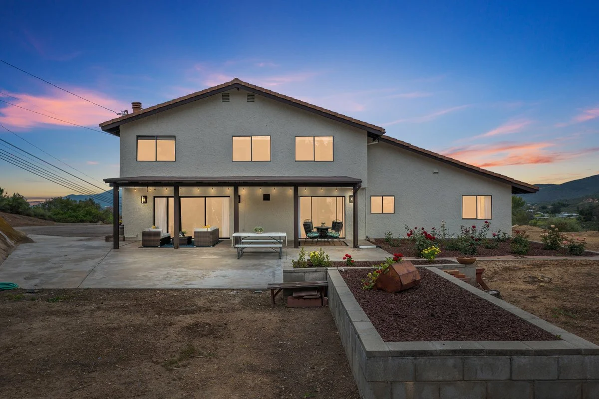 A two-story house with a white stucco exterior, large windows, and a brown tiled roof, viewed during sunset, with a patio area and flower beds in the foreground.