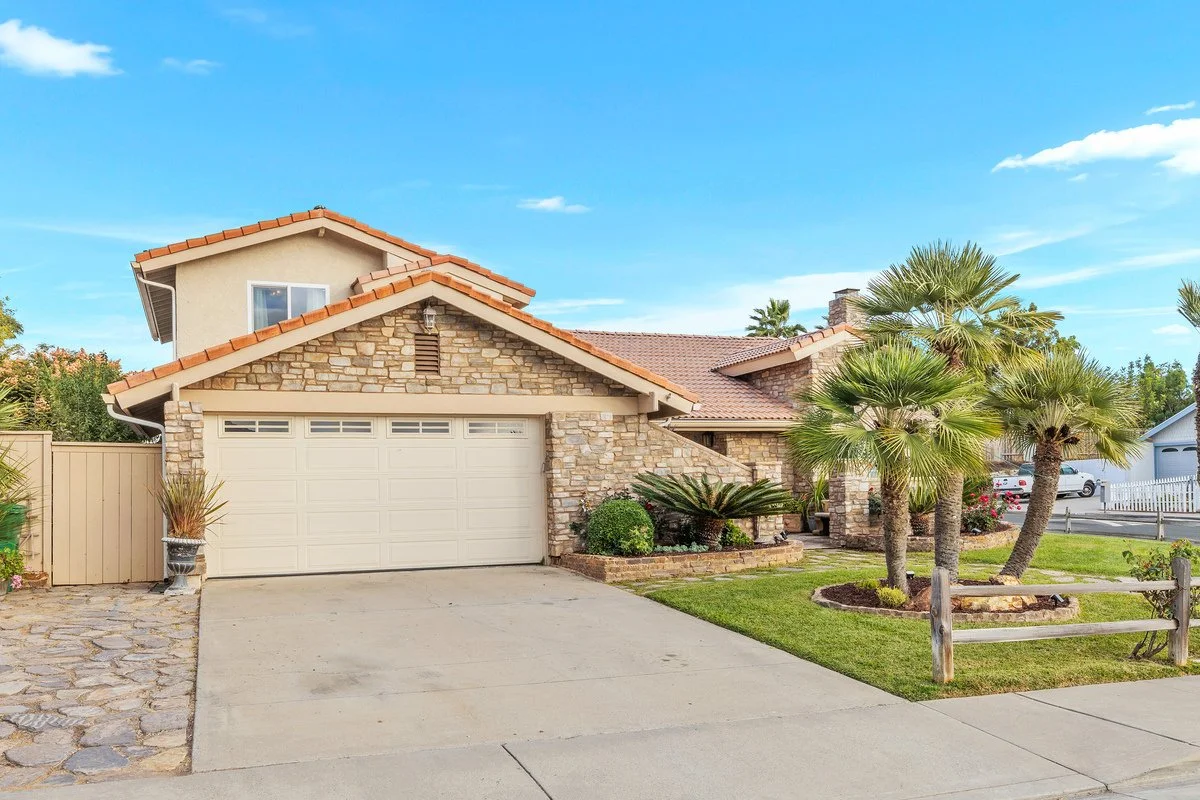A suburban house with a two-car garage, stone exterior, and a front yard with palm trees and green grass under a blue sky.