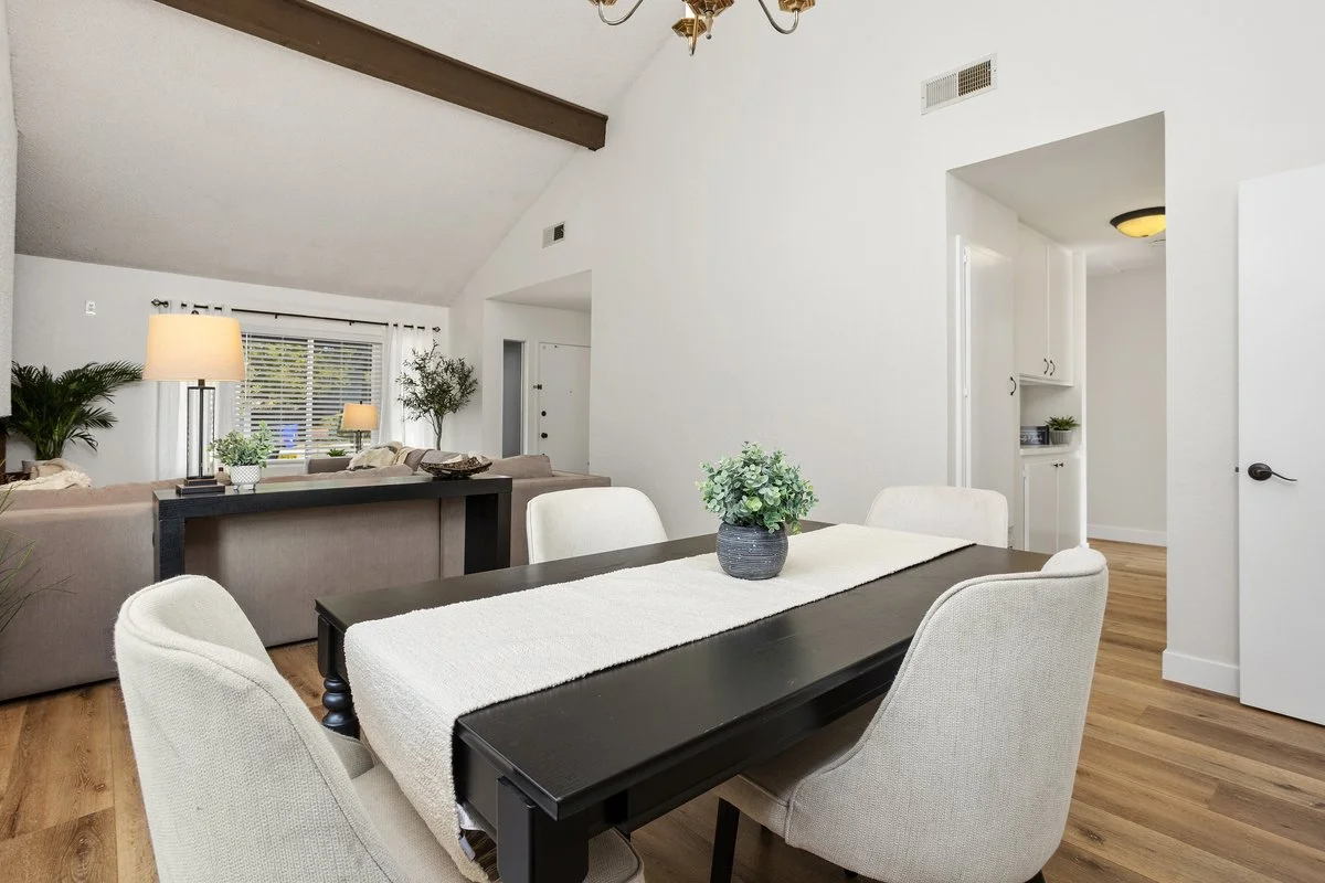 A dining room with a black table and four beige upholstered chairs, a white table runner, and a potted green plant. In the background, a living area with beige couches, lamps, and large windows with blinds.