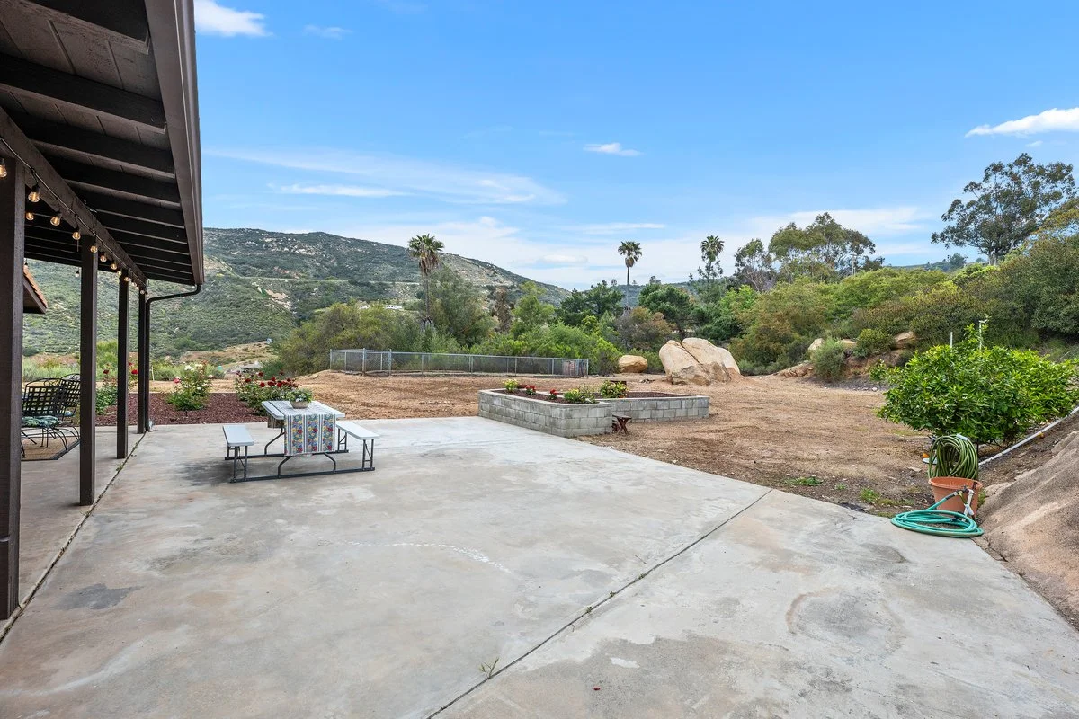Backyard patio with concrete slab, garden bed with red flowers, and hillside with trees and rocks under a blue sky.