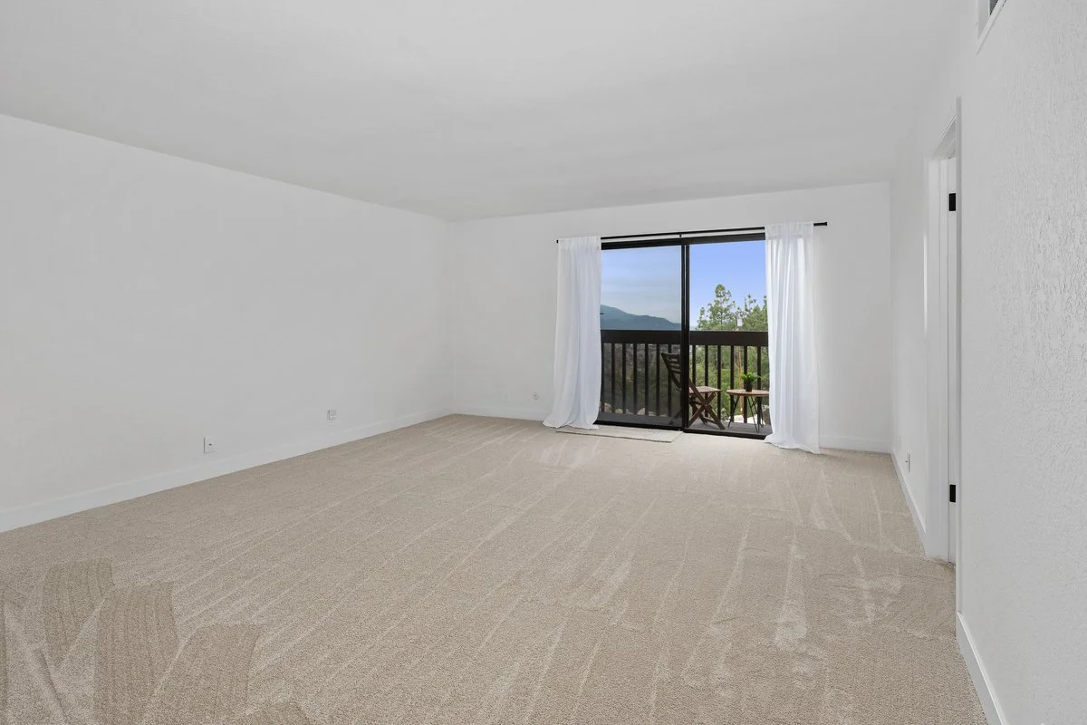 Empty living room with white walls, beige carpet, sliding glass door with white curtains, and small balcony with outdoor furniture. Mountains and trees visible outside.
