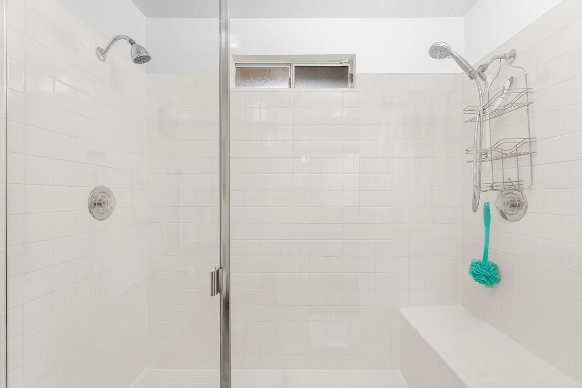 Empty shower with white tiles, two showerheads, a small window, a built-in bench, and a shower caddy with a scrubber hanging from one of the showerheads.