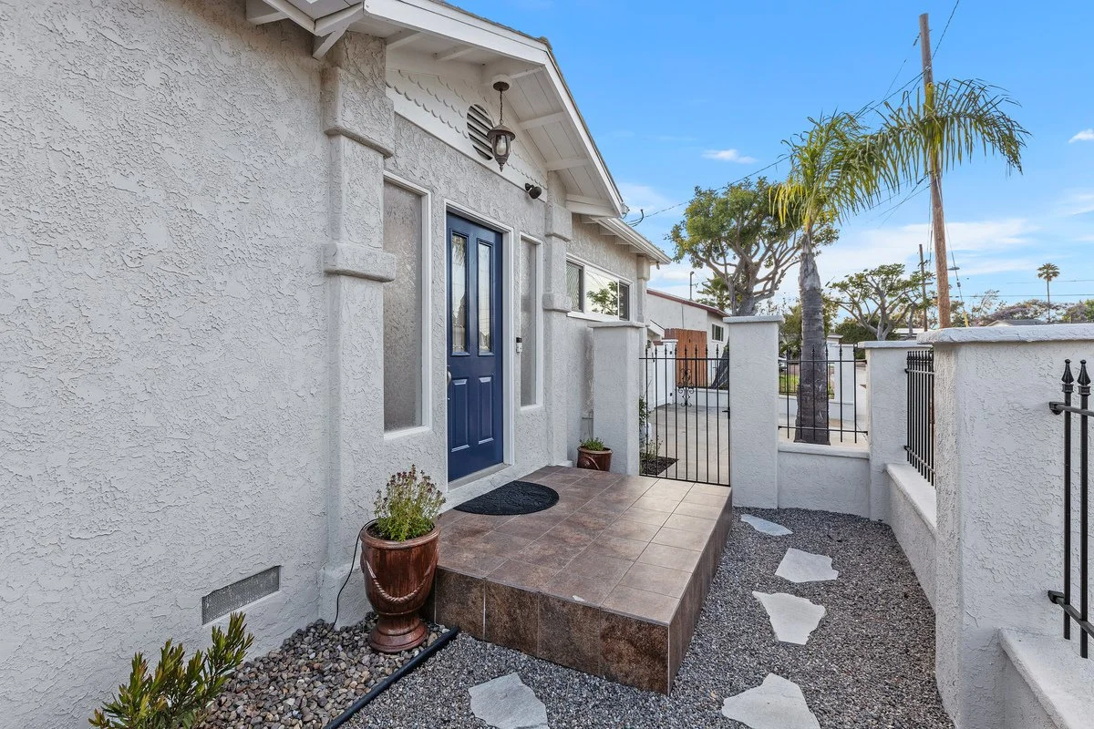 Front porch with a blue door, potted plant, tiled steps, and a small pathway with stepping stones, surrounded by a white stucco wall and fence, under a clear blue sky with trees in the background.