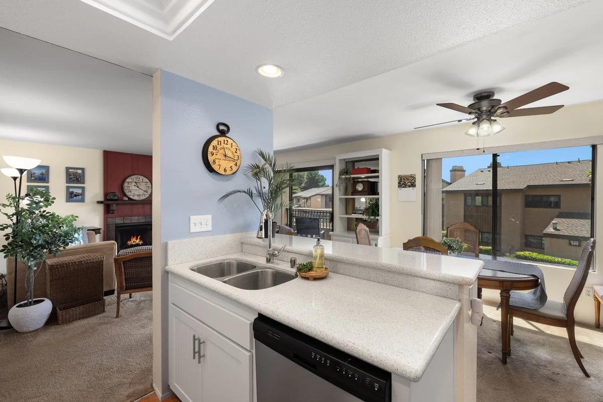 Open concept living space view from kitchen, showing dining area with a table and chairs, sliding glass door, sitting area with armchair, fireplace, and wall decor, with a ceiling fan and large windows letting in natural light.
