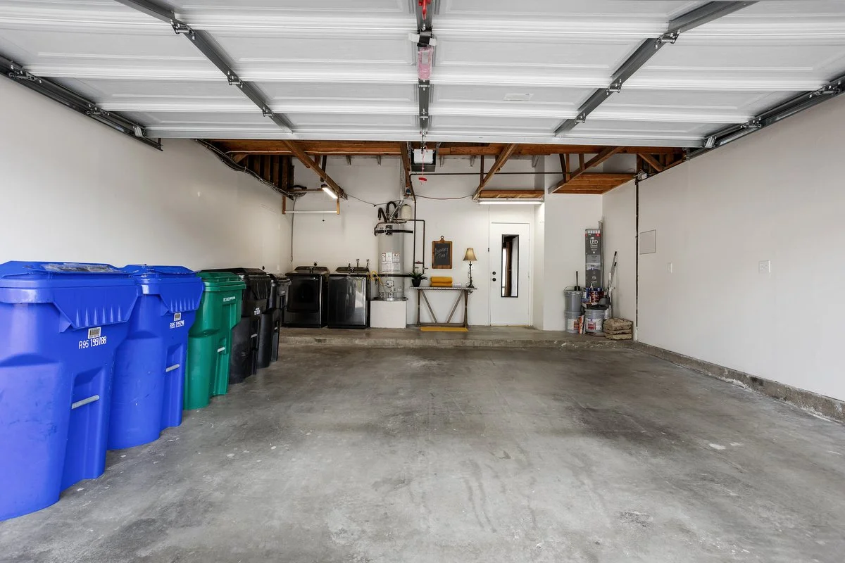 Empty garage with laundry machines, recycling bins, and cleaning supplies, featuring concrete floors, white walls, and an open garage door.