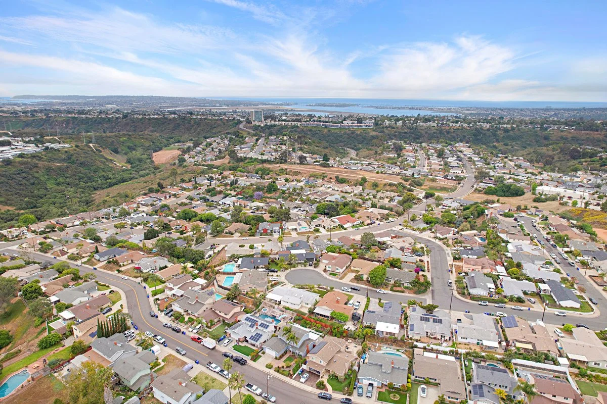 Aerial view of a suburban neighborhood with houses, streets, trees, and a distant bay or ocean under partly cloudy skies.