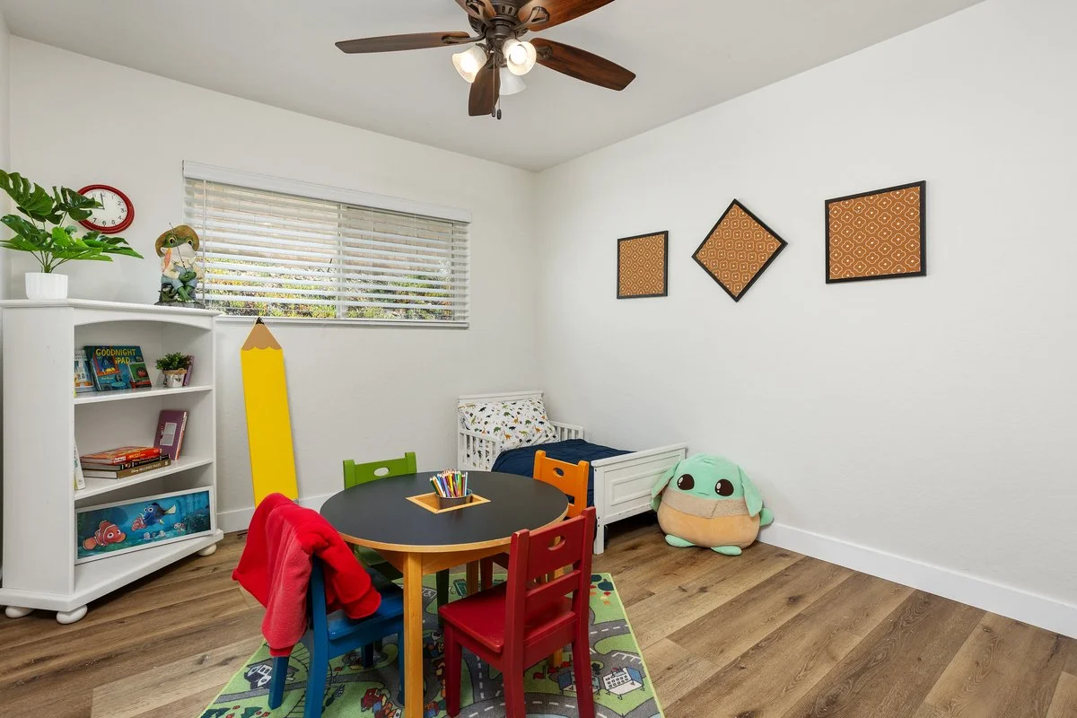 A children's bedroom with a white crib, plush turtle toy, a small round table with colorful chairs, a bookshelf with children’s books, a yellow toy sword, a window with blinds, and wall art with three geometric shapes. The room has wood laminate floo