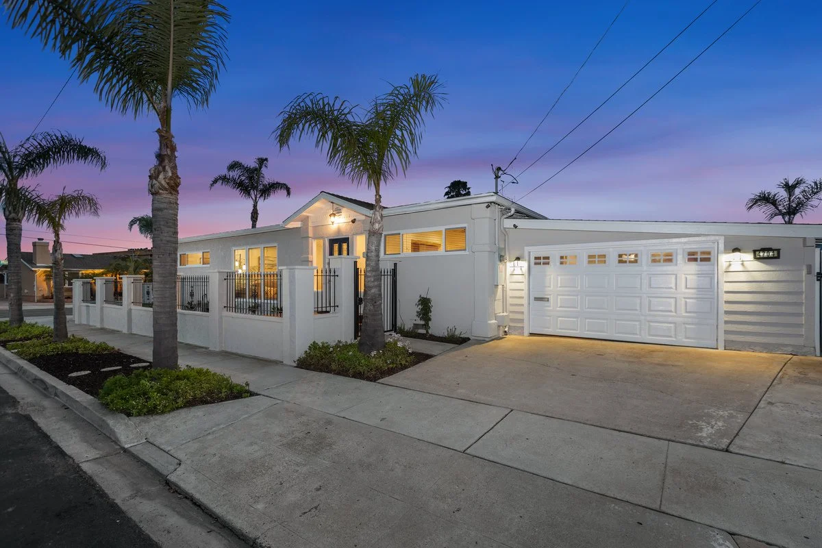A modern white house with a gated front yard and palm trees, illuminated at dusk with a purple and blue sky in the background.