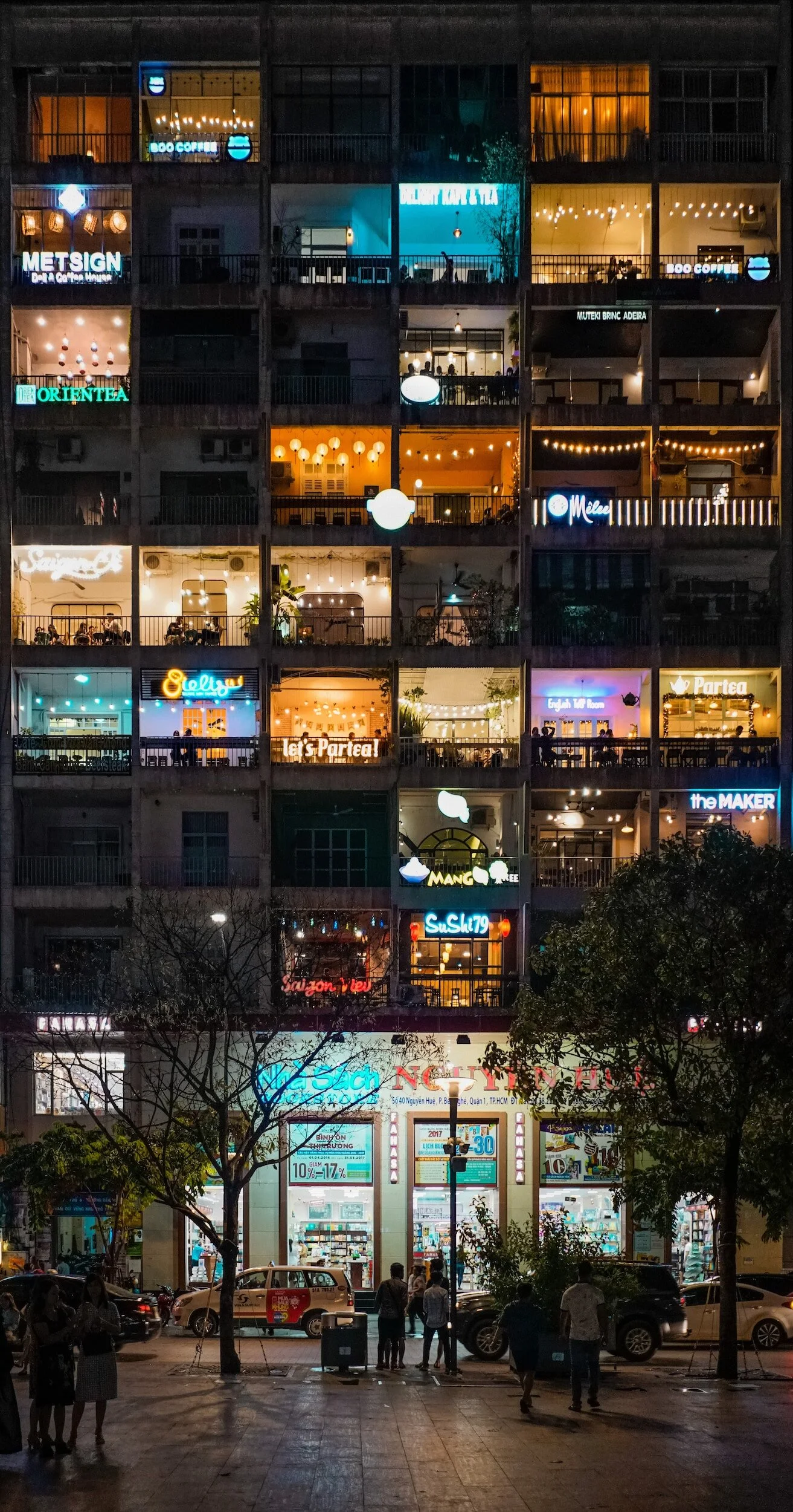 A multi-story building with lit-up balconies decorated with string lights and neon signs, viewed at night. The ground level features a brightly lit storefront with people walking and cars parked outside.