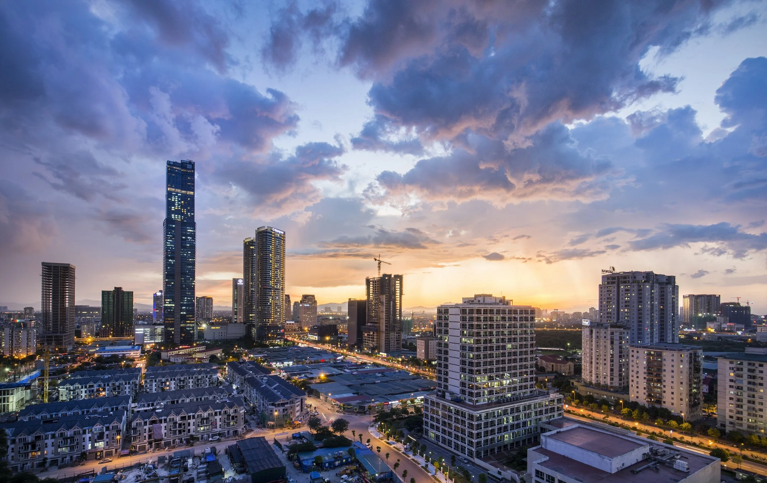 City skyline at sunset with high-rise buildings, clouds, and streaks of sunlight.