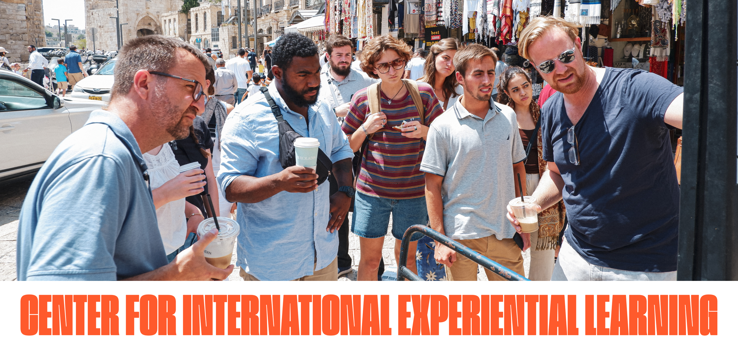 Group of diverse young adults standing in line outdoors, some holding drinks, near a street market with shops and hanging textiles in the background.