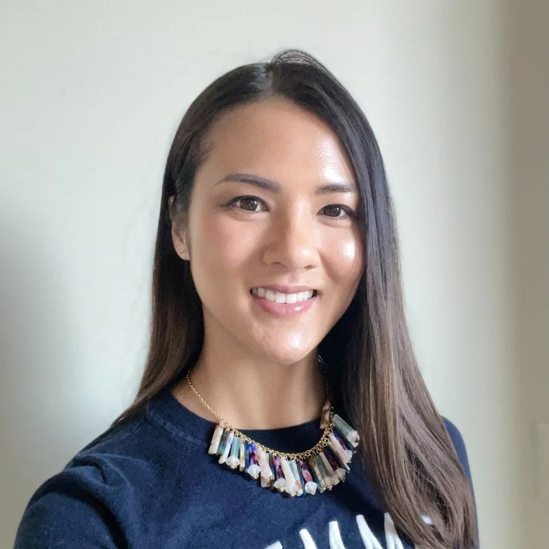 A woman with long dark hair smiling at the camera, wearing a dark shirt and a colorful statement necklace against a plain background.