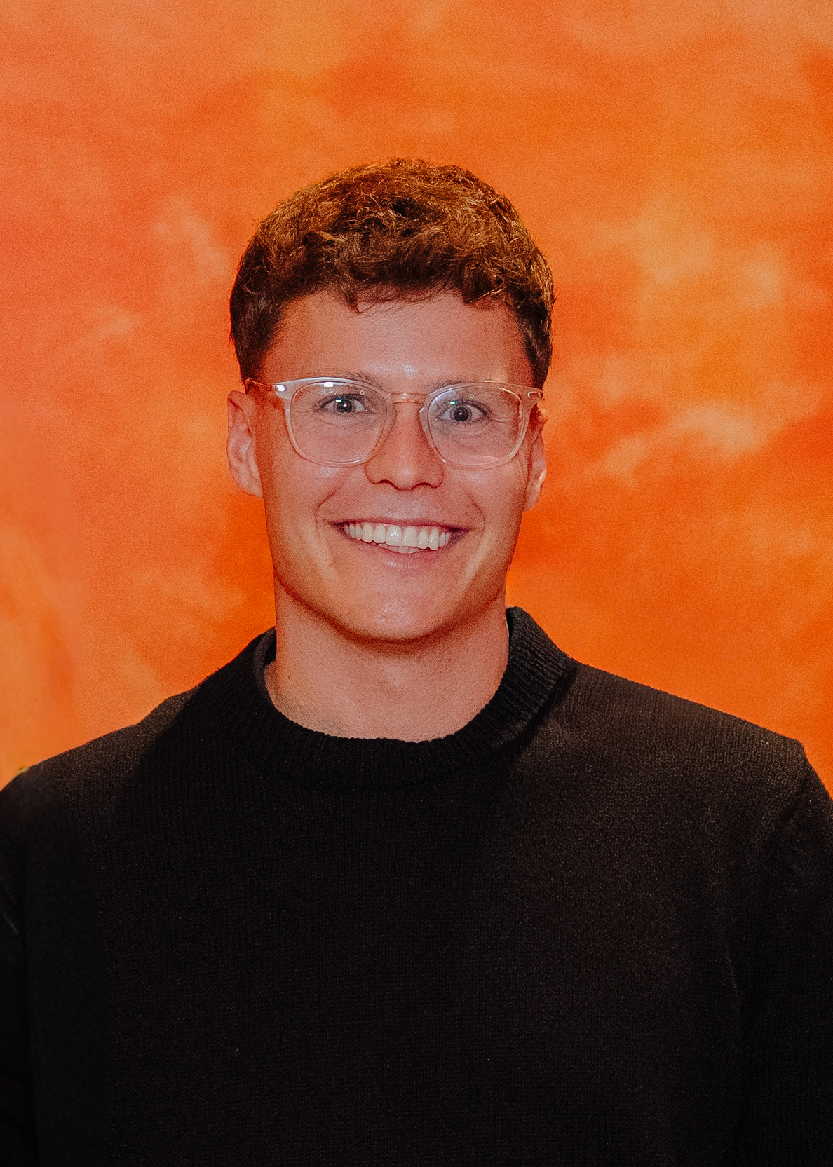 A young person with short curly hair, glasses, and a broad smile, wearing a black sweater, standing against an orange textured background.