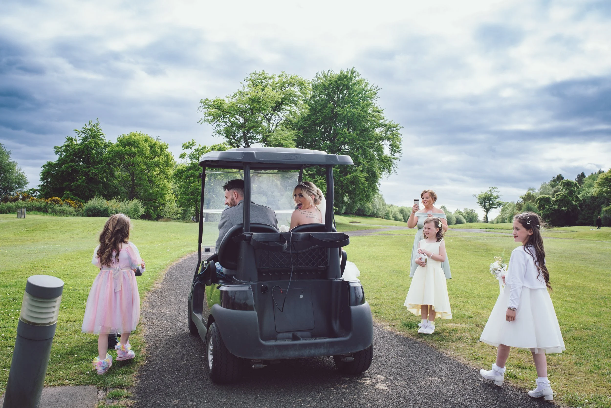 Bride and groom travelling together in a golf buggy with flower girls following them