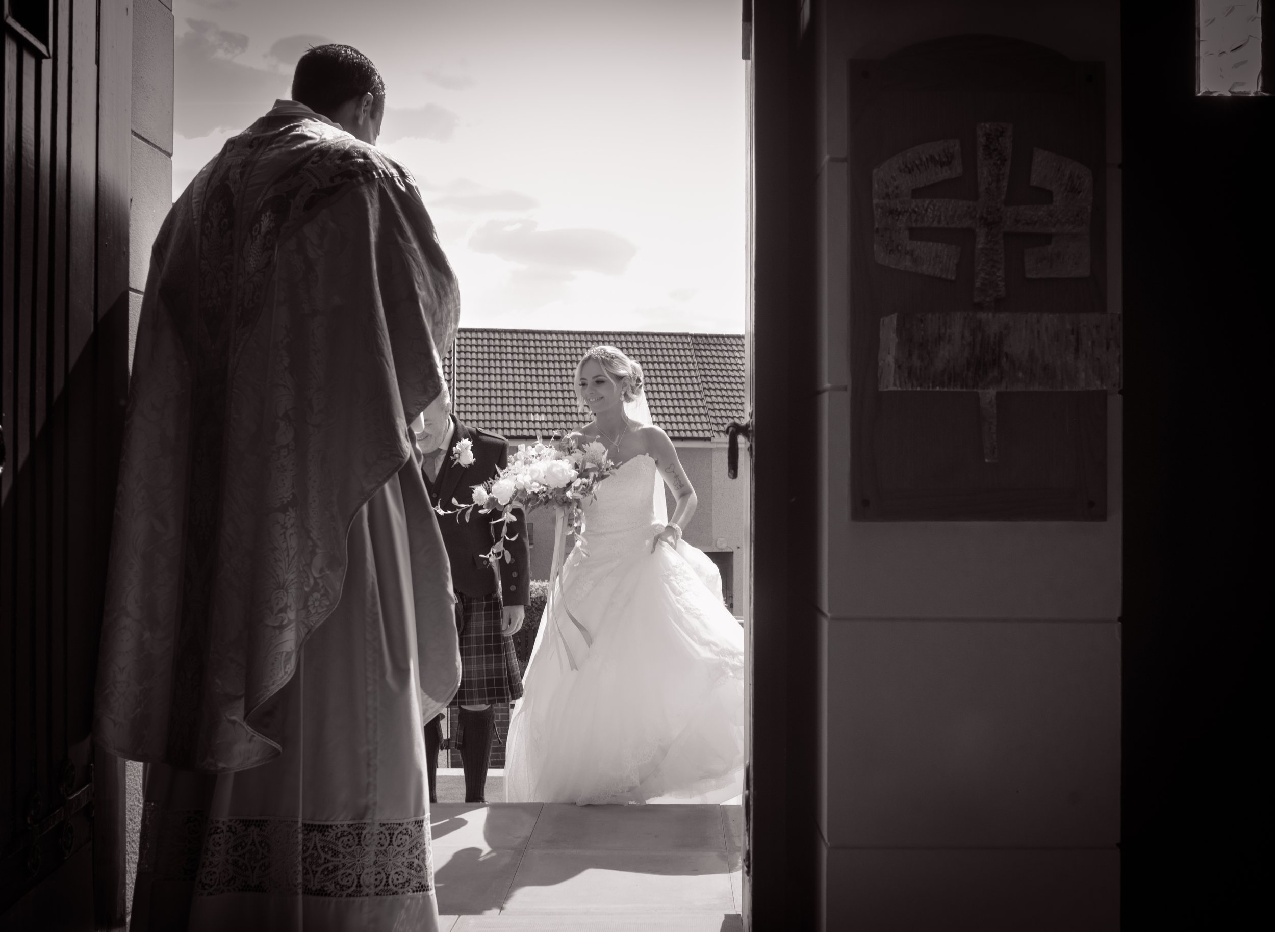 Bride arriving at her wedding ceremony with a piper and priest welcoming her at the church entrance