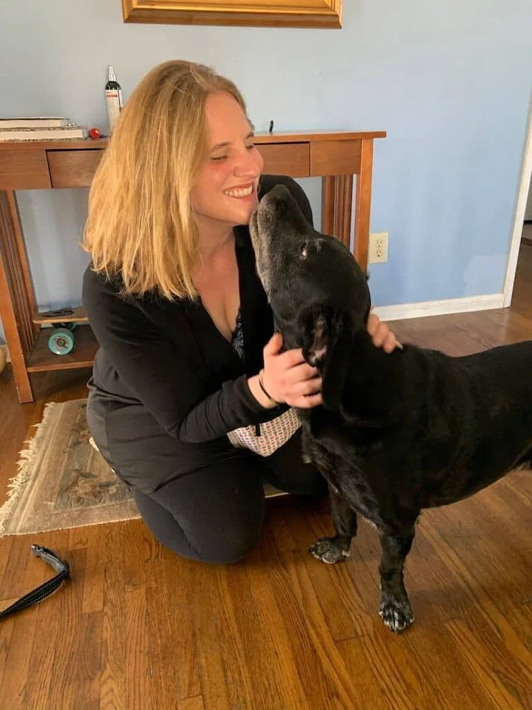 A woman kneeling on a hardwood floor, smiling with her eyes closed, as she interacts affectionately with a large black dog. The woman has shoulder-length blonde hair and is wearing a black top and pants. In the background, there is a wooden table, a