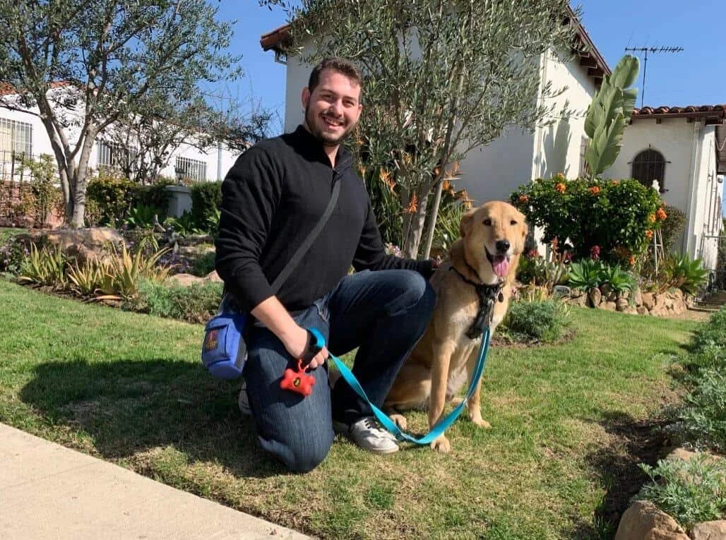 A man kneeling on grass with a leash around his new dog, both smiling, in a sunny garden with plants and a house in the background.