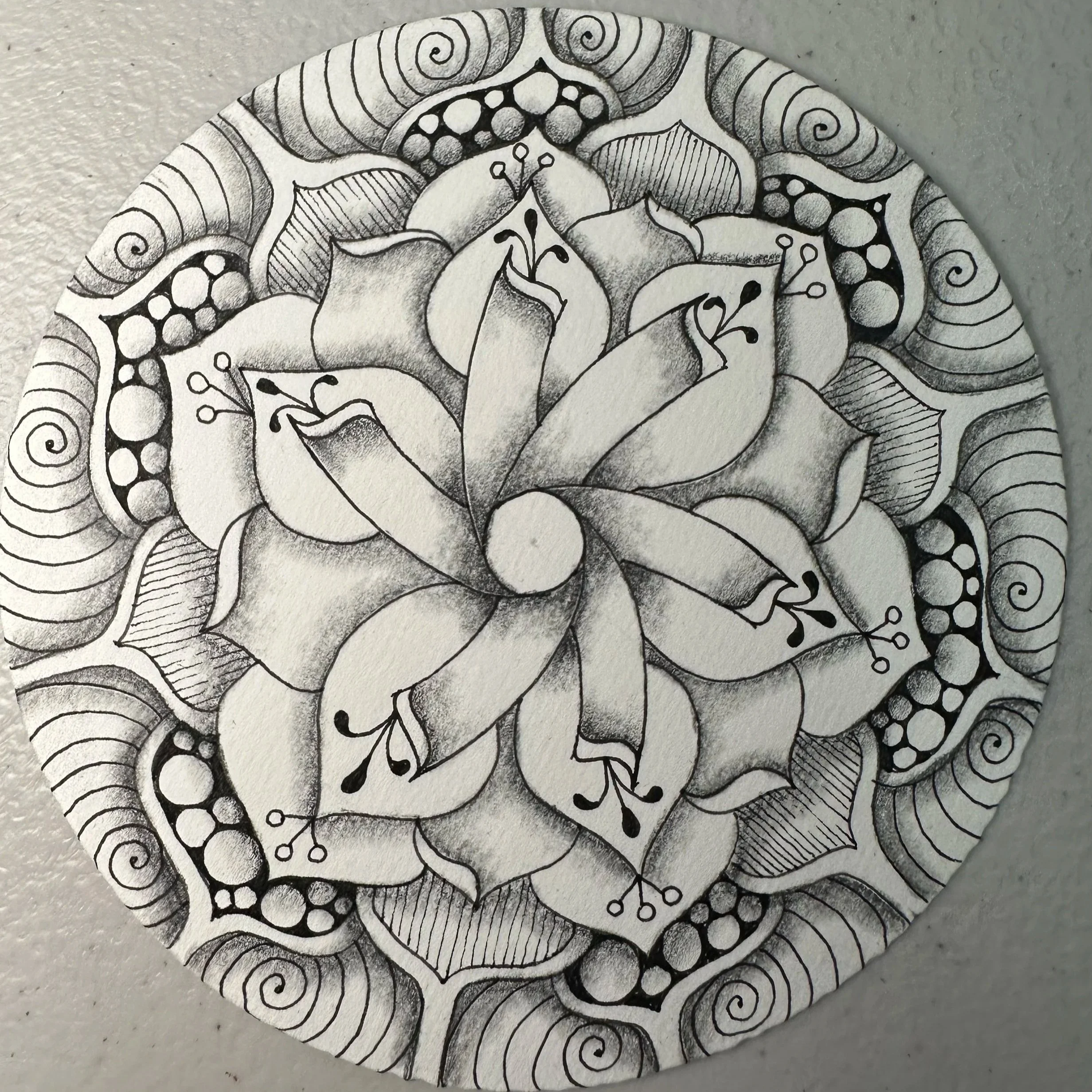 Black and white ink drawing of a floral mandala with symmetrical petals, leaves, and intricate patterns radiating outward.