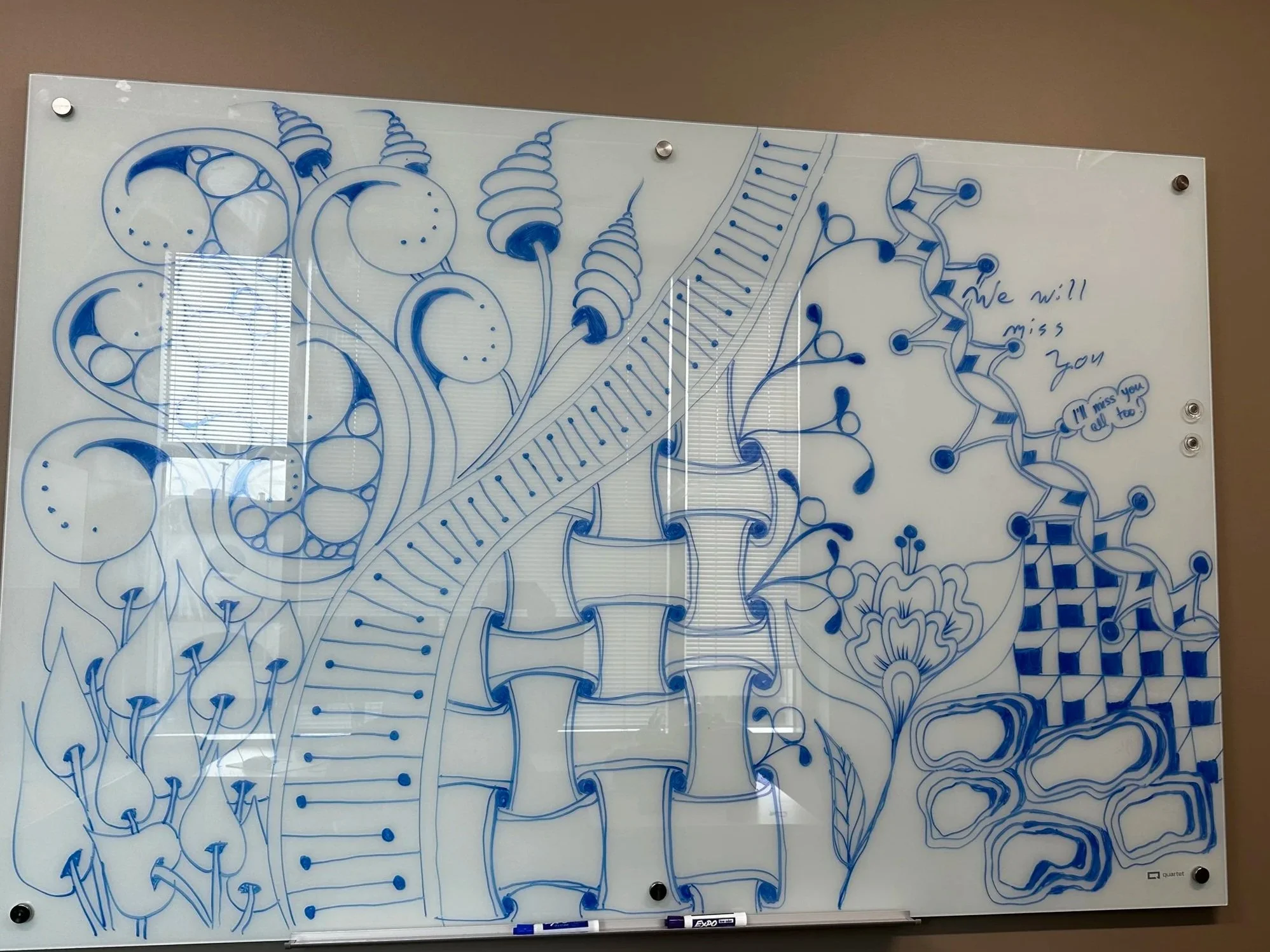 Blue marker drawing on a whiteboard featuring abstract and floral patterns, with the words "We will miss you" and "I'll miss you all too" written in cursive.