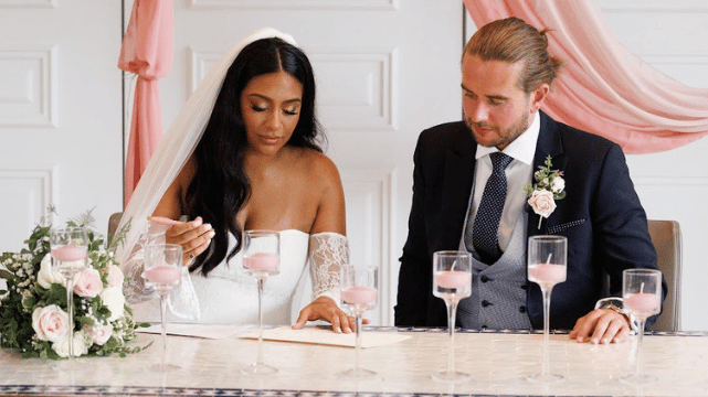 Bride signs a document at a table with pink candles, while groom in a suit sits beside her; both look focused and elegant.