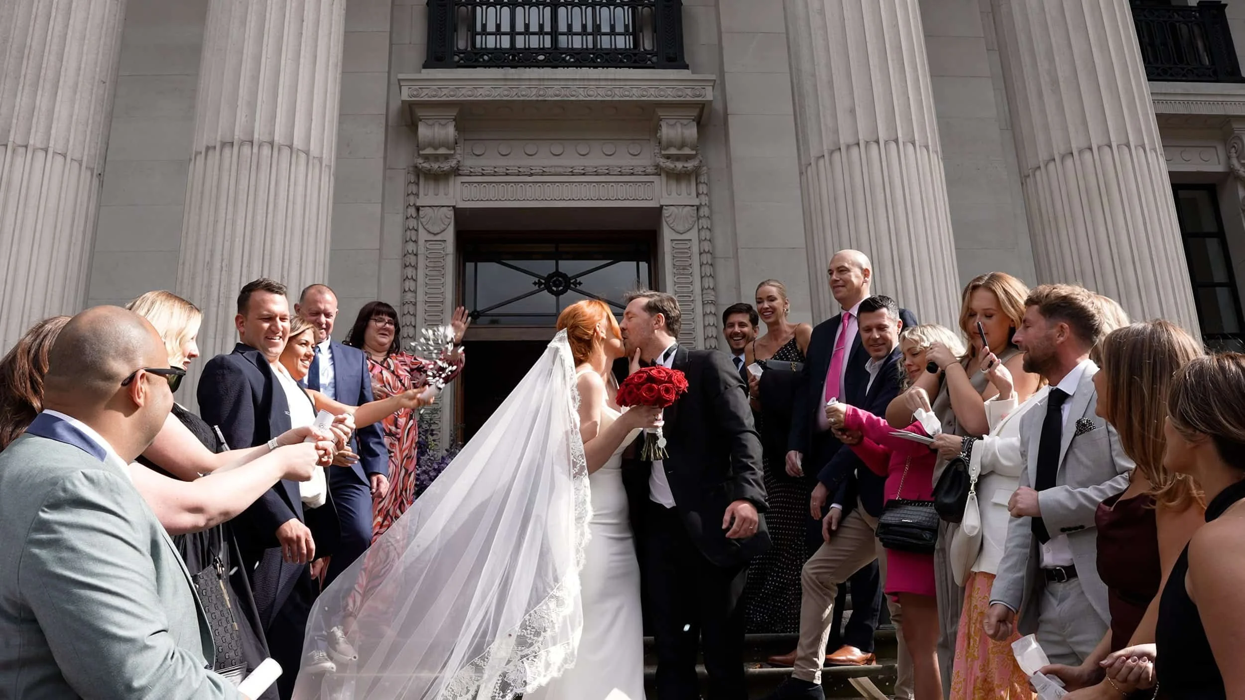 couple kiss on wedding day on steps of Old Marylebone Town Hall London surrounded by family