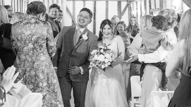 A bride and groom smile, walking down the aisle together as wedding guests applaud and take photos in a bright, rustic venue.