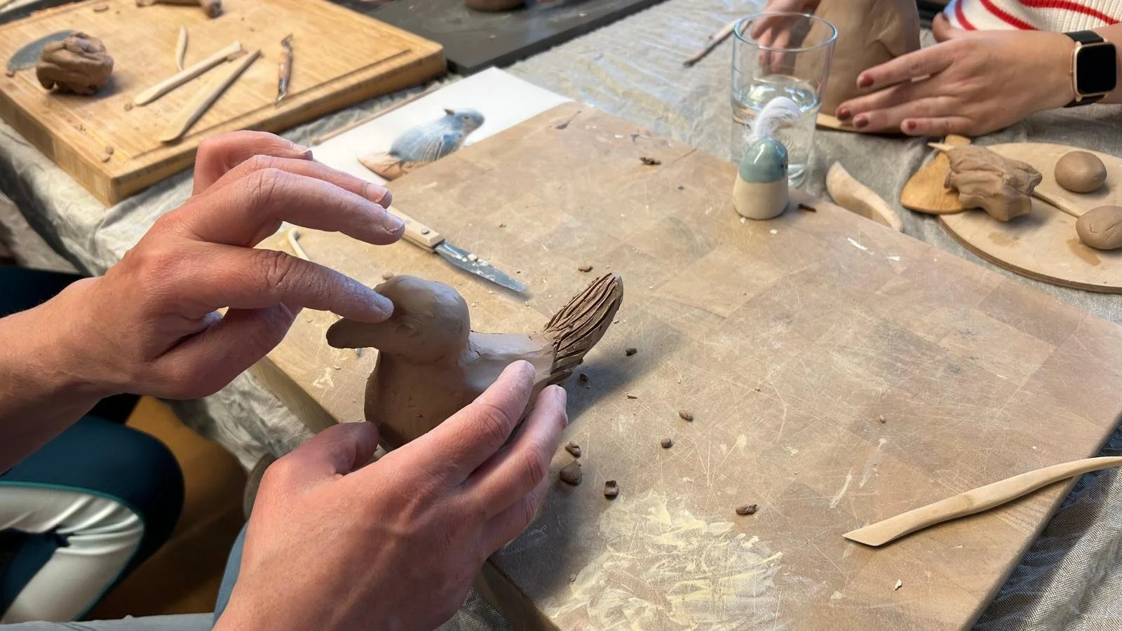 Personen modellieren eine kleine Vogelfigur aus Ton auf einem Tisch, umgeben von Werkzeugen und weiteren Tonarbeiten.