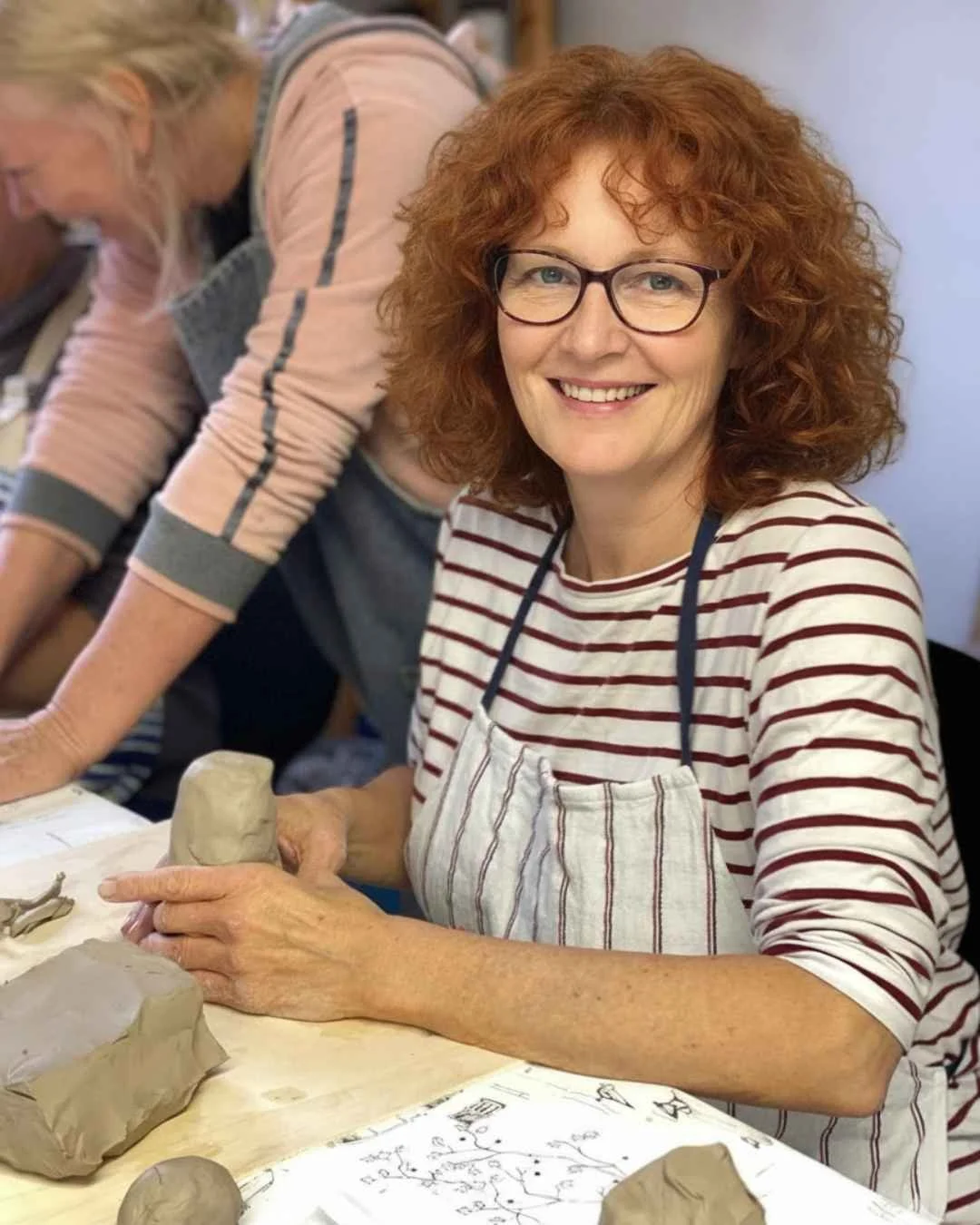 Frau mit roten, lockigen Haaren und Brille, die bei einem Keramik-Treppen-Workshop töpfert. Sie trägt ein weißes Hemd mit roten Streifen und eine Schürze, während sie an einem Tisch mit Tonarbeiten arbeitet.