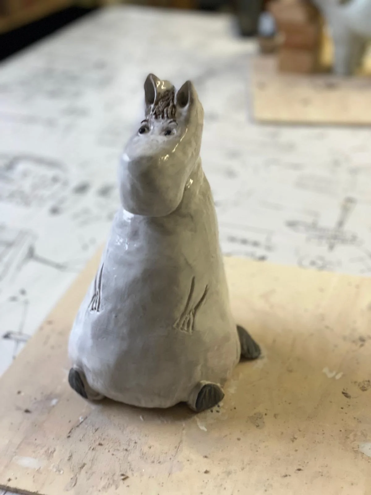 Eine Tier-Skulptur, die wie ein Nilpferd aussieht, aber mit einem Tierkopf im Dinosaurierstil und einem kurzen Körper. Es steht auf einem Holztisch in einem Raum, vermutlich in einer Werkstatt oder einem Studio. Der Hintergrund ist unscharf.
