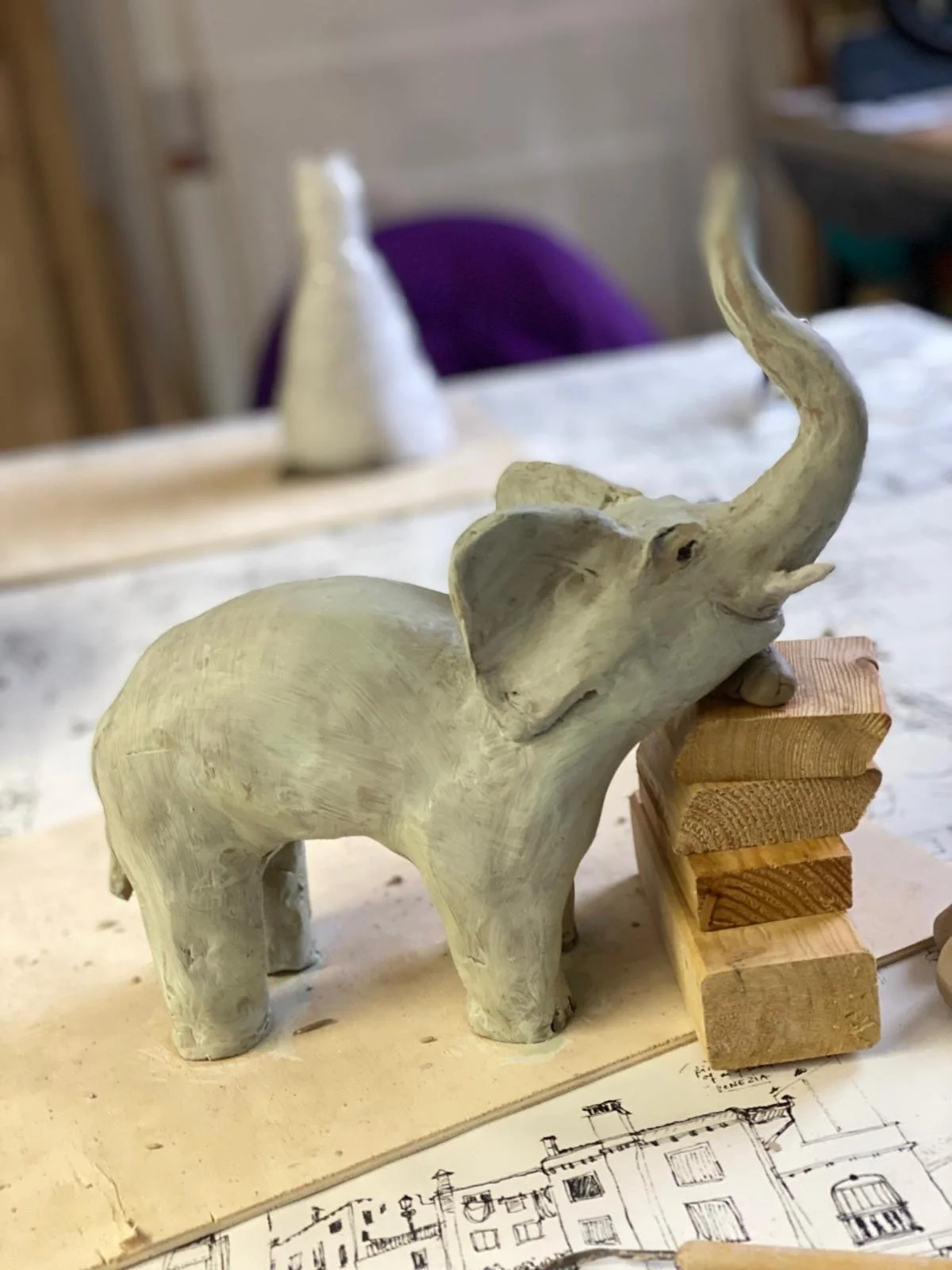Ein handgeschnitzter Elefanten-Baum Skulptur aus Holz, auf einem Tisch mit Baustoffen und Bauplänen im Hintergrund.