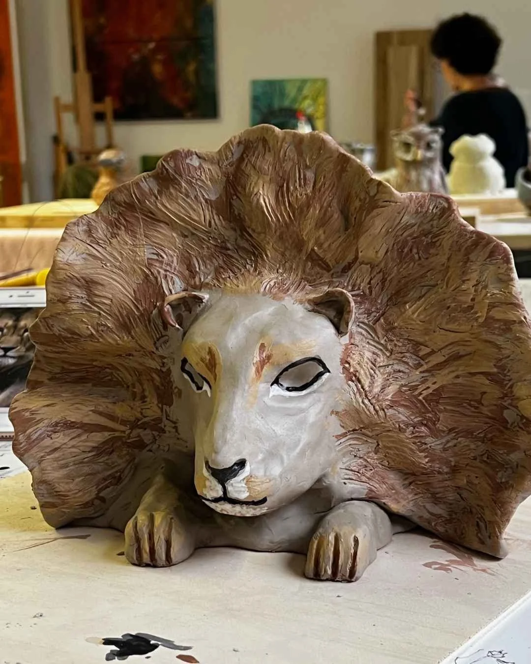 Skulptur eines Löwenkopfes aus Holz, mit einem detaillierten, realistischen Gesicht und großen, kräftigen Mähne. Im Hintergrund sind Menschen und Kunstwerke in einem Atelier sichtbar.