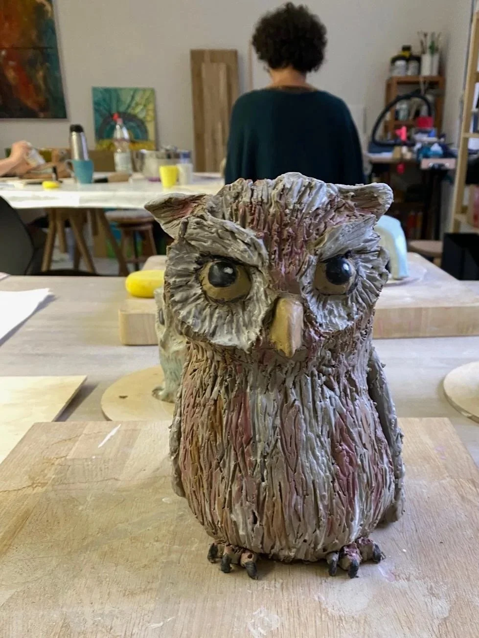 Eine detaillierte Keramikskulptur einer Eule auf einem Holzuntergrund, im Hintergrund ein kreatives Atelier mit einer Person, die an einem Tisch arbeitet.