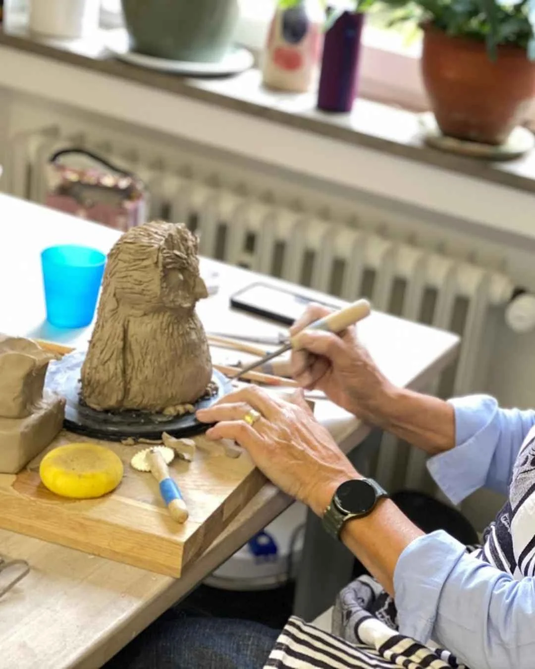 Eine ältere Person modelliert eine Skulptur eines Löwen aus Ton auf einem Tisch. Im Hintergrund sind Fenster, Pflanzen und Dekorationsgegenstände sichtbar.