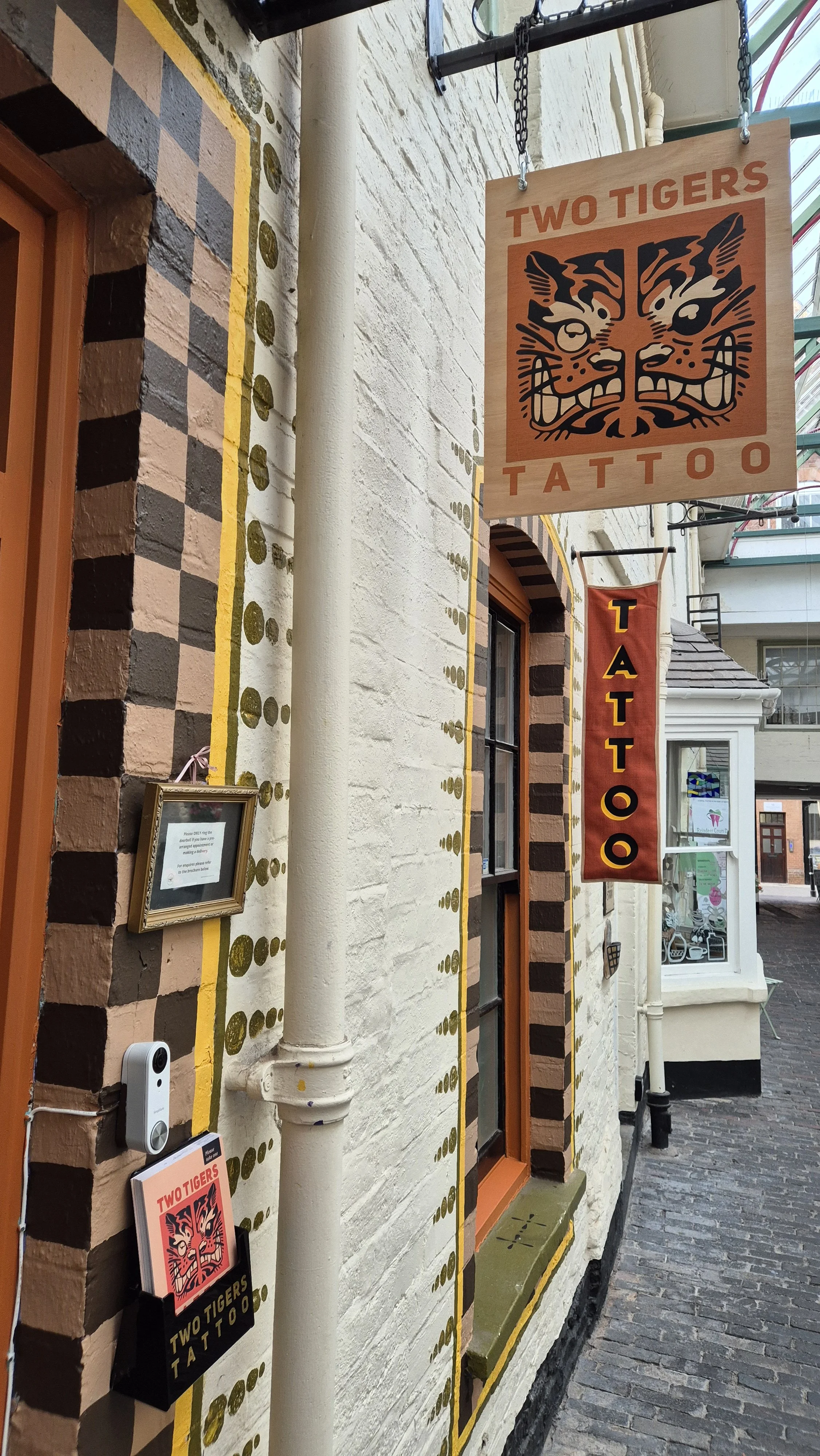 Exterior of a tattoo shop called Two Tigers Tattoo with signs hanging outside, featuring a tiger face logo and decorative brickwork on the building.
